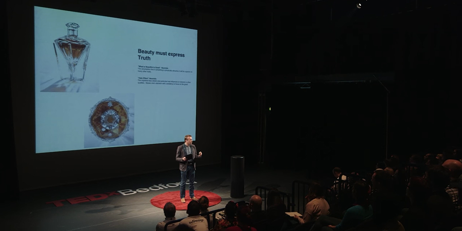 Jeremy Lindley sobre el escenario de las charlas TEDxBedford. Una pantalla de fondo proyecta una hermosa botella de cristal con whisky.