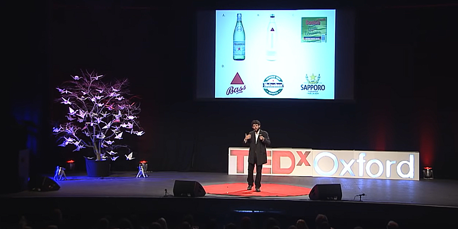 Alejandro Salgado Montejo dando una charla en el escenario de TEDxOxford. De fondo se proyecta una imagen con varios logos y botellas de cervezas reconocidas.