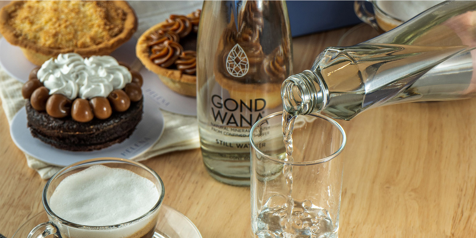 Una botella de agua mineral premium Gond Wana se vierte en un vaso delante de otra botella de Gond Wana, sobre una mesa servida con pastelería dulce, diseño de packaging por Tridimage.
