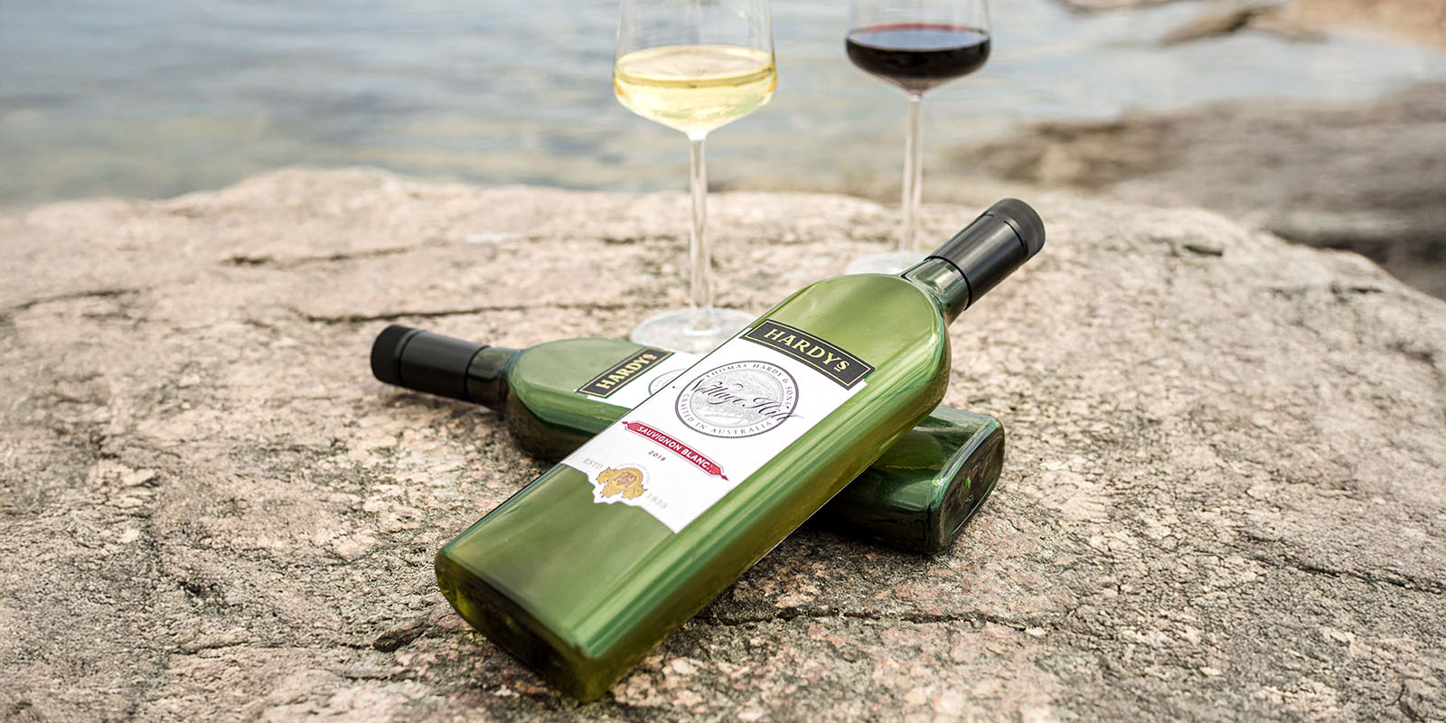 Botellas de vino planas de PET reciclado Garçon Wines en un entorno natural: tres botellas de vino Hardy’s de diseño plano y ecológico, hechas de PET 100% reciclado, junto a dos copas con vino blanco y tinto. Están colocadas sobre una superficie rocosa cerca de un cuerpo de agua, resaltando la sostenibilidad del packaging.
