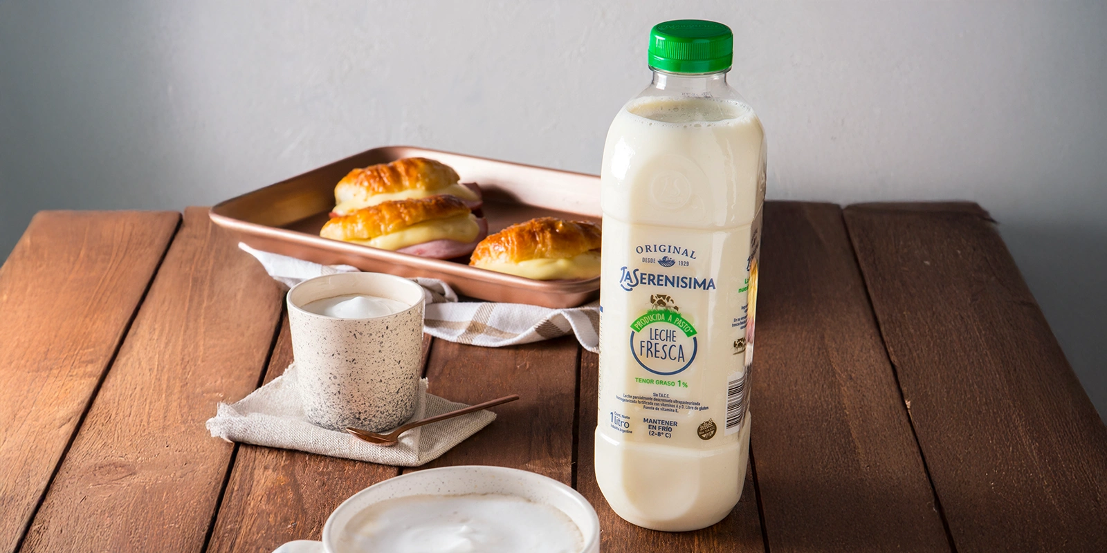 Botella de Leche Fresca La Serenísima en un desayuno con medialunas y café con leche espumosa.