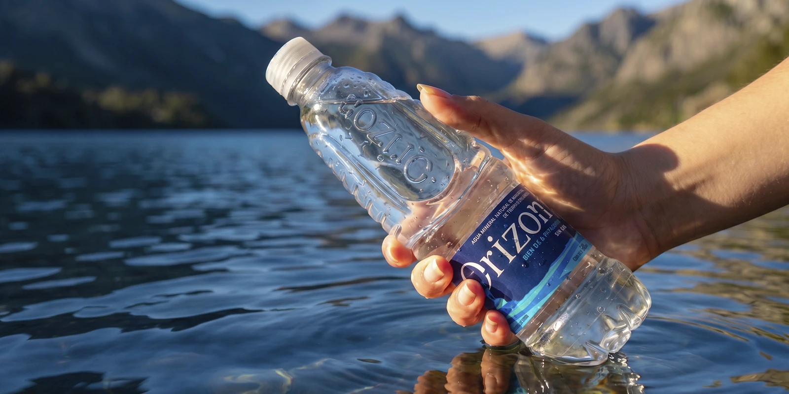Mano sosteniendo una botella de agua Orizon frente a un lago y montañas de la Patagonia argentina al atardecer.