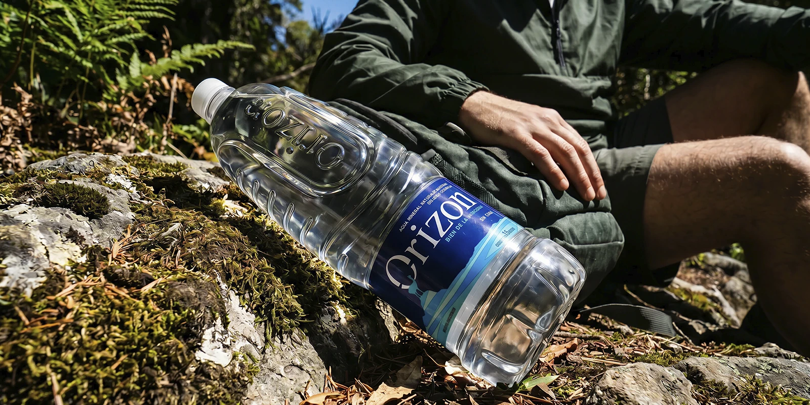 Botella de agua mineral Orizon de 1.5 litros descansando sobre una roca con musgo junto a un senderista.