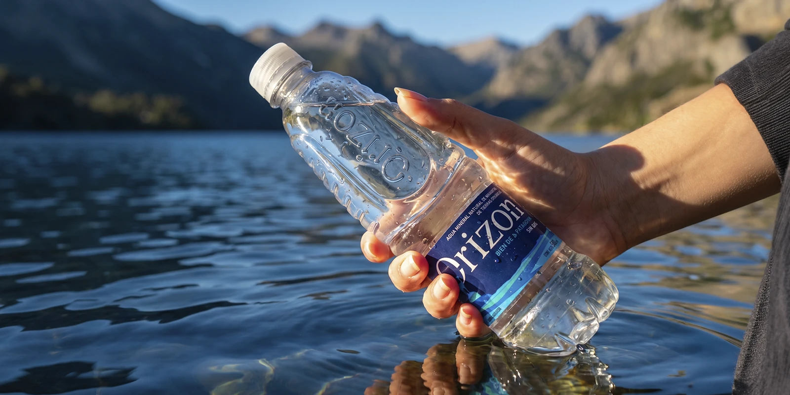 Mano sosteniendo una botella de agua Orizon frente a un lago y montañas de la Patagonia argentina al atardecer.