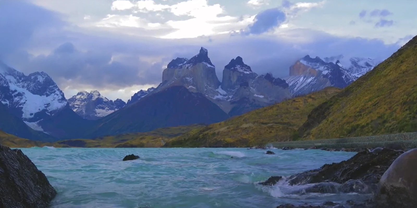 Paisaje de la Patagonia con montañas nevadas, cielo nublado y un lago de agua turquesa con oleaje sutil.